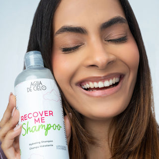 A smiling woman with closed eyes holds a bottle of AGUA DE CIELO Recover Me sulfate free Shampoo next to her face against a light gray background.