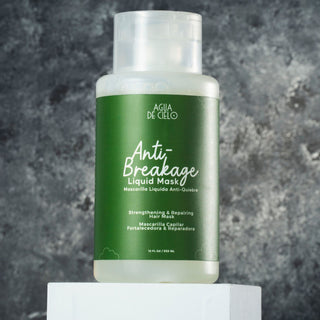 A bottle of AGUA DE CIELO Anti-Breakage Liquid Mask hair mask, featuring a green label and clear cap, displayed on a white pedestal against a textured gray background.