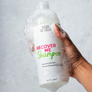 A hand holding a clear plastic bottle of Agua de Cielo Recover Me Shampoo, the best hydrating sulfate free paraben free shampoo, covered in white foam, against a light gray textured background.