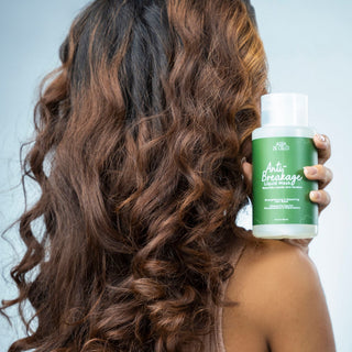 A woman with long, wavy brown hair is shown from the back, holding a bottle of breakage repair Anti Breakage liquid mask against a light gray background.