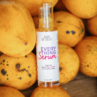 A bottle of Agua de Cielo Everything Serum stands upright in the center, surrounded by a pile of yellow mangoes. The bottle has a clear cap and a white label with purple and pink text.
