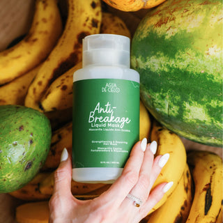 A hand with white manicured nails holds a bottle of Agua DE Cielo Anti Breakage Liquid Mask, a hair breakage repair treatment, surrounded by fresh fruits including bananas, a watermelon, and a green avocado.