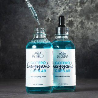 Two glass dropper bottles of Agua de Cielo hair energizing drops filled with bright blue liquid, set against a dark textured gray background with one bottle open and a glass pipette above it.
