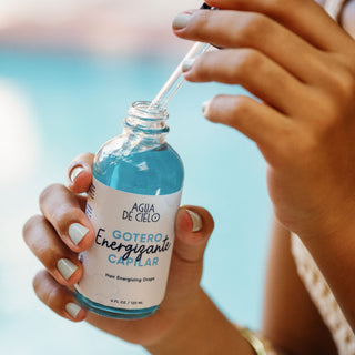 A person holds a small glass bottle of hair follicule stimulator with a white label reading 'AGUA DE CIELO GOTERO Energizante CAPILAR' against a blurred outdoor background.