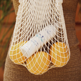 A white mesh produce bag hanging on a brown burlap carrier, filled with yellow citrus fruits and a white bottle of olive oil. Includes hair heat protectant for hair by agua de cielo