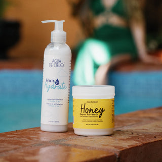 Two hair care products from Agua de Cielo standing on a wooden ledge by a pool: a white pump bottle labeled Hair 24 Hydrate and a white jar with a yellow label labeled Honey Repair Pre-poo.