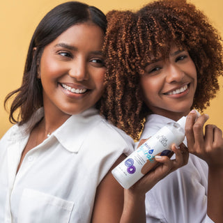 Two smiling women standing close together and holding a white bottle of hair product against a warm yellow studio background.