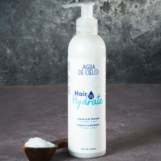 A white pump bottle of Hair Hydrate leave-in conditioner by Agua de Cielo, displayed on a dark textured surface with a small wooden spoon of white product in the foreground.