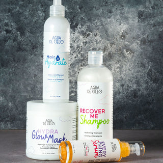 Agua de Cielo Hair Recovery Kit arranged on a dark textured background: a tall Hair Hydrate spray bottle, a Recover Me Shampoo bottle, a Hydra Glow Mask jar, and Everything Serum spray bottle lying on its side.