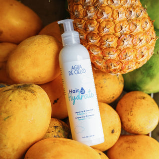 A bottle of Agua de Cielo Hair Hydrate heat protectant and leave in conditioner placed among yellow mangoes and a pineapple, with a green mango in the background.