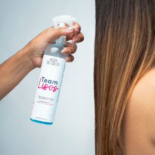 A hand holding a spray bottle of Agua DE CIELO Team Lisos heat protection spray for straightening hair, shown spraying onto the hair of a person with long brown hair, against a light gray background.