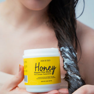 A person holds a white jar labeled 'Honey Repair Pre-Poo' by Agua de Cielo in front of their shoulder, with dark hair coated in a white creamy product hanging beside it.