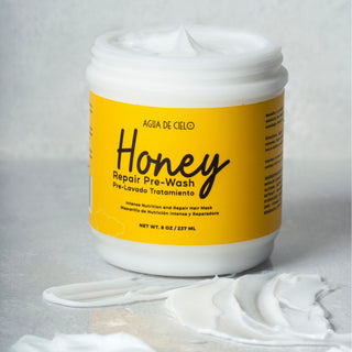 A white plastic jar of Agua DE CIELO Honey Repair Pre-Shampoo treatment for damaged hair mask with a bright yellow label, set on a light gray textured surface with white cream smeared in front.