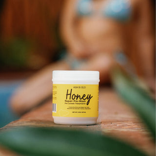 A jar of AGUA DE CIELO Honey Repair Prepoo pre-lavage treatment sits on a wooden surface with a blurred background of a person and green leaves.