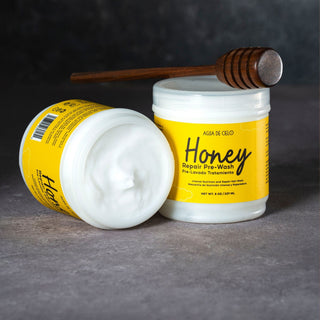 Two jars of honey repair pre-poo with a wooden honey dipper resting on top, set against a dark gray textured background.