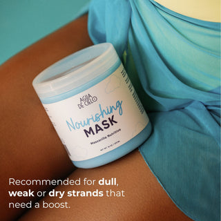 A jar of Agua de Cielo Nourishing hair mask for dry hair lies on a person's shoulder with turquoise fabric draped nearby. The label reads 'Nourishing MASK' and includes a text callout about dull, weak, or dry strands needing a boost.