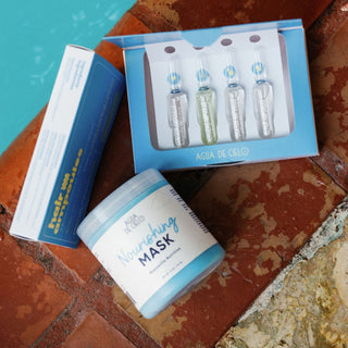 Agua de Cielo featuring a jar of Nourishing Mask, four glass ampoules in a blue box, and an additional blue box, arranged on a rustic terracotta tile surface next to a turquoise pool.
