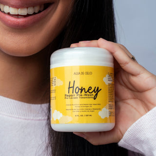 A close-up of a person holding a white jar of 'Agua de Cielo' Honey Repair Pre-champu against a light gray background.