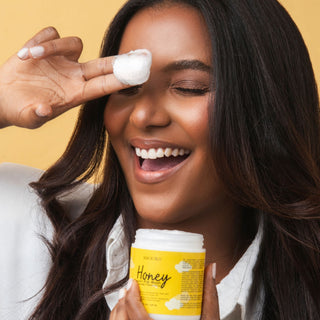 A woman with long dark hair smiles while applying white cream from a jar labeled 'Honey preshampoo treatment' to her eye. She is holding the yellow and white jar against a solid yellow background.