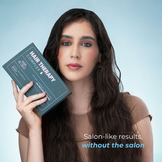 A woman with long dark brown hair holds a dark teal-green box labeled 'HAIR THERAPY COMPLEX 5 professional style hair ampoules' by the brand 'Agua de Cielo'. She is wearing a brown ribbed top against a light blue gradient background, with the text 'Salon-like results, without the salon' in the bottom right corner.