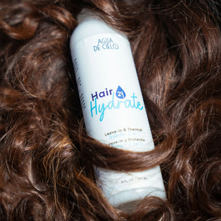 A white bottle of Hair Hydrate leave-in conditioner by AGUA DE CIELO, wrapped in and surrounded by voluminous, glossy brown hair. The product label features blue and purple text.
