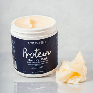A white plastic jar of Agua de Cielo Protein Therapy Har Mask for curly hair with a navy blue label, shown open with cream-colored mask visible inside and a dollop of the mask placed beside it on a light gray reflective surface.
