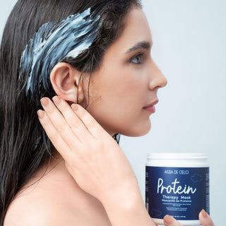 A woman with dark hair applies a white hair mask to her scalp, holding a white jar of AGIA DE CIELO Protein Therapy Hair Mask in the foreground against a light gray background.