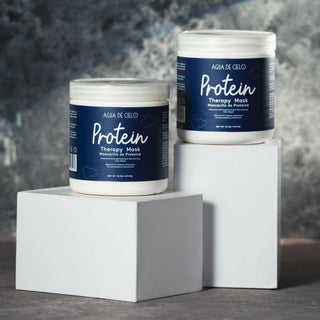 Two white plastic jars of agua de cielo protein therapy mask with dark blue labels, displayed on white pedestals against a textured gray background.