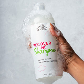 A hand holding a bottle of Agua de Cielo Recover Me Shampoo, covered in white foam bubbles, against a light gray textured background.