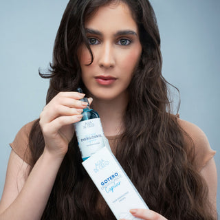 A woman with long, wavy brown hair holds a bottle of Agua de Cielo scalp stimulator. She is wearing a beige short-sleeve top against a solid light blue-gray studio background.