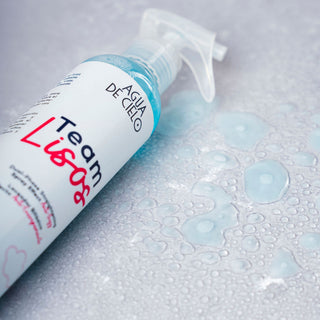 A spray bottle of Aqua de Cielo Team Lisos hair straightening spray, lying diagonally on a wet, water-droplet-covered surface.